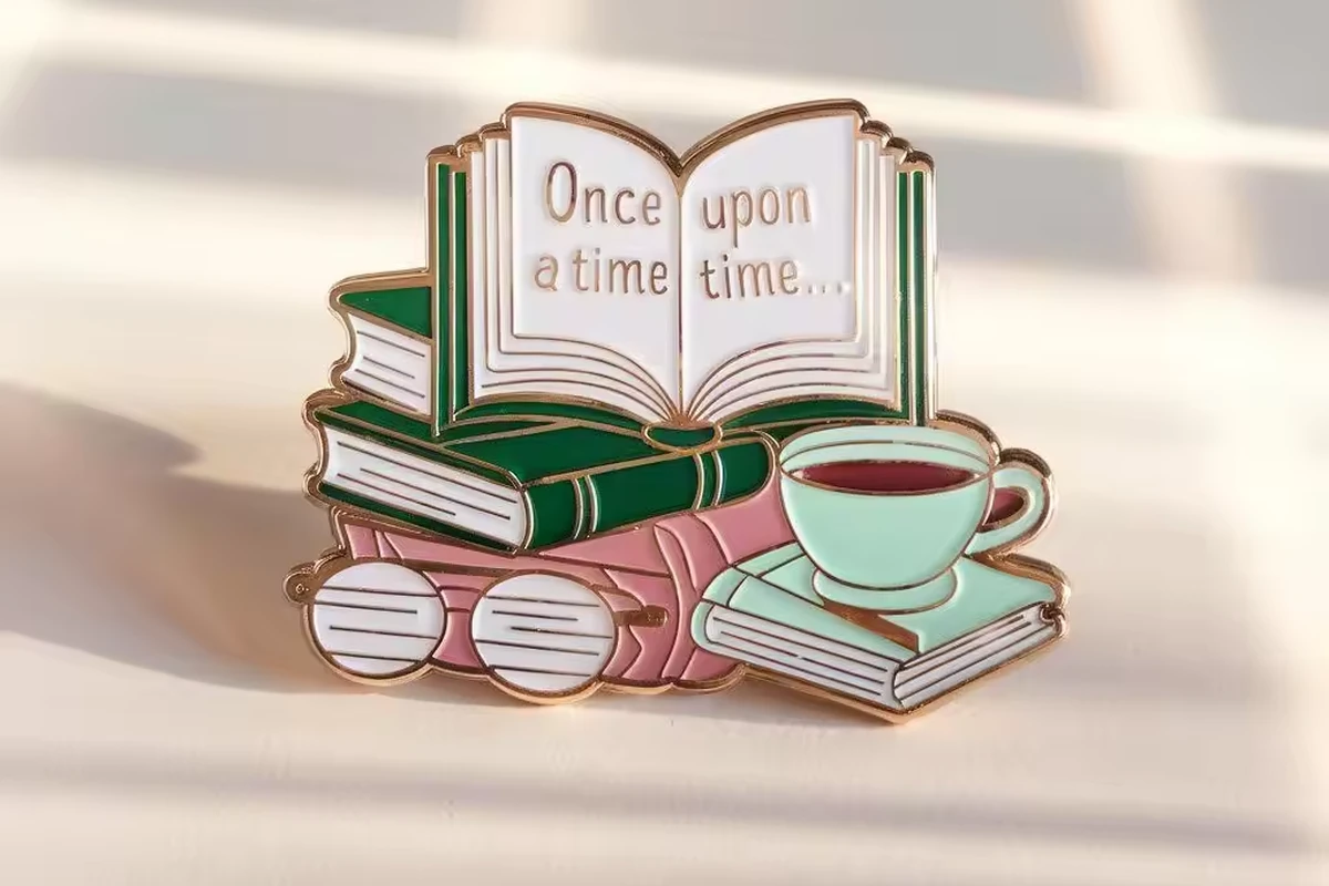 A book, glasses and teacup-shaped enamel pin on softly lit cream fabric.