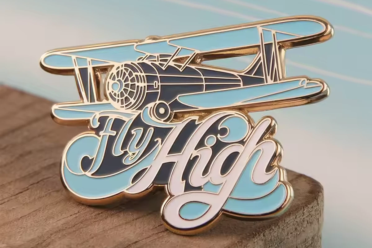 A blue and gold biplane pin with the words Fly High, displayed on a wood surface.