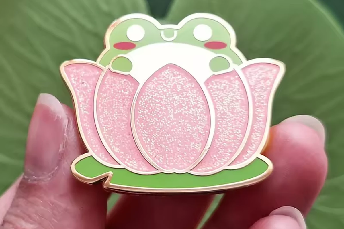 A hand holding a green frog pin nestled inside a glittery pink lotus flower.