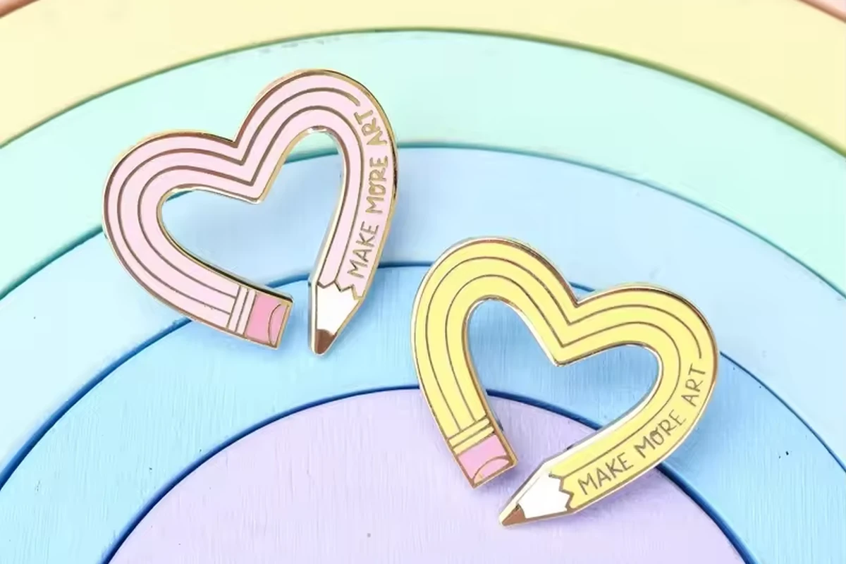Pink and yellow heart-shaped pencil pins with Make More Art lettering on a pastel rainbow background.