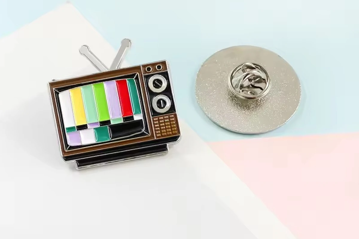 A retro television-shaped pin with colored screen bars beside a round pin back fitted with a butterfly clutch.