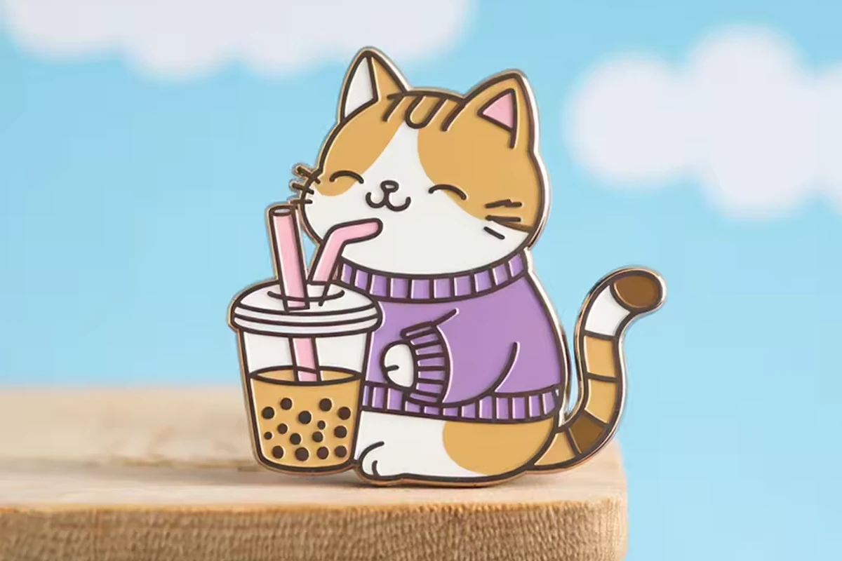 An orange cat enamel pin holding bubble tea on a wooden stand against a blue cloud background.