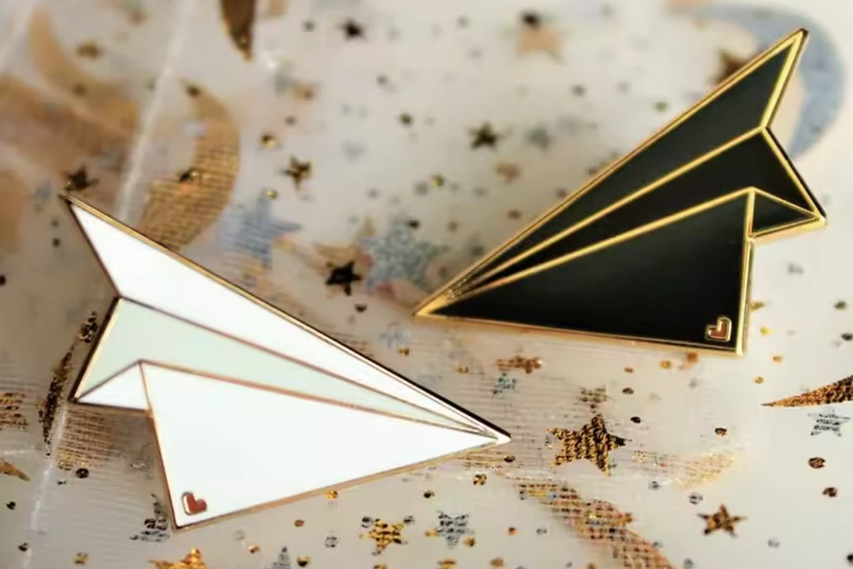 Black and white paper-airplane enamel pins with gold-colored outlines on patterned fabric.