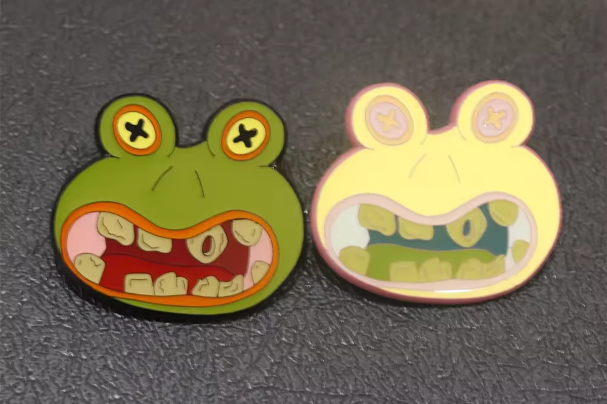 Two frog-shaped enamel pins in green and pale yellow on a dark textured surface.