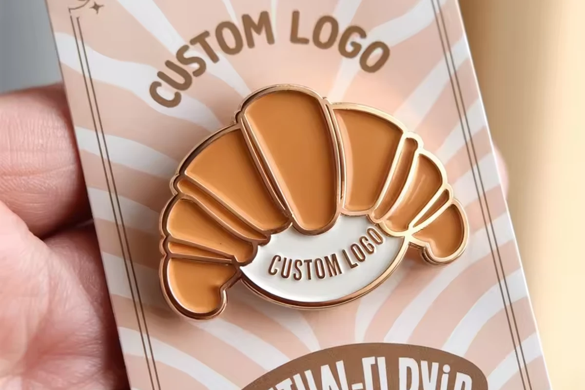 A hand holding a croissant-shaped pin on a pink and white printed backing card.