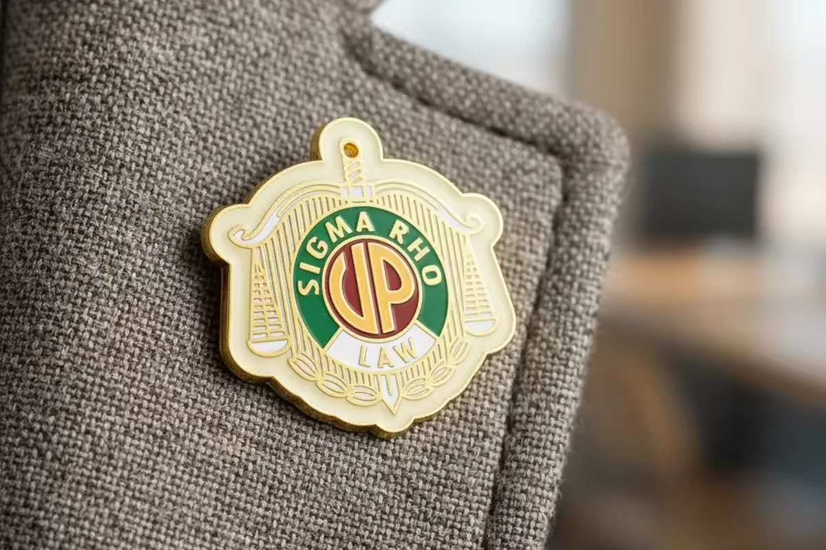 A gold, cream and green fraternity-style crest pin worn on a gray jacket.