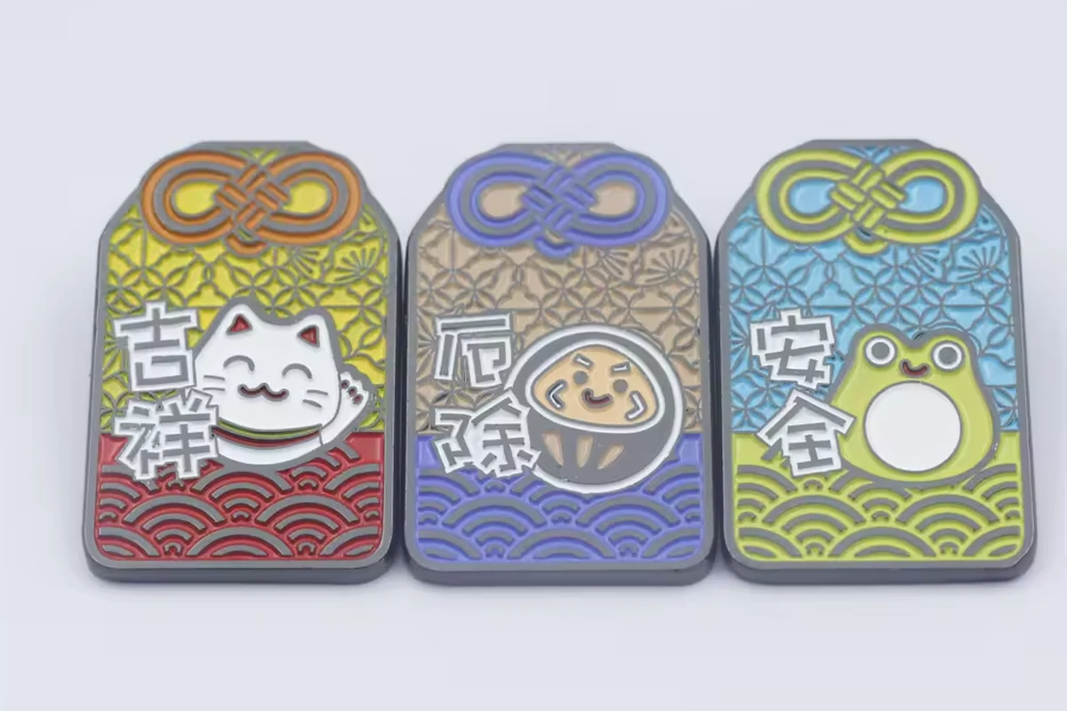 Three colorful rectangular charm pins with cat, rice-ball and frog designs on a pale background.