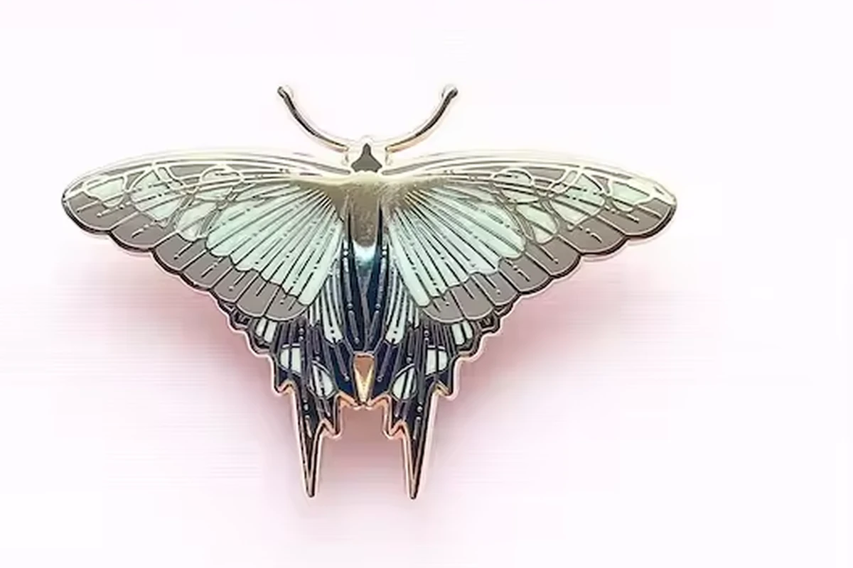 A pale green and blue moth-shaped metal pin with detailed wings on a soft pink background.