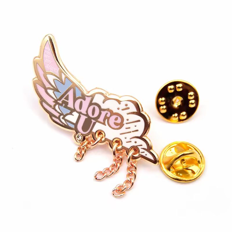 Pastel pink, blue and white wing-shaped pin with Adore U lettering, three dangling chains and a small clear stone, beside two gold-tone butterfly clutches on white.