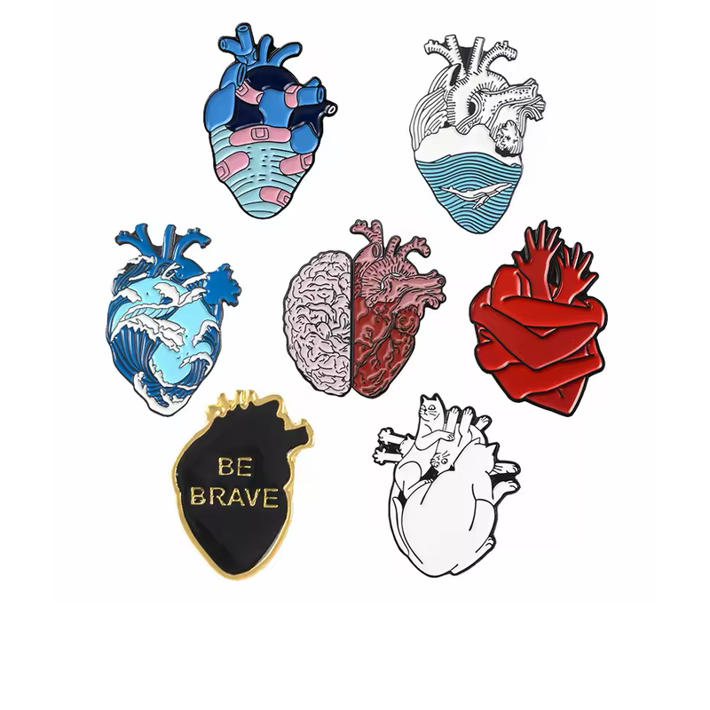 Seven anatomical heart-themed pins with bandages, ocean waves, a brain-and-heart design, red folds, BE BRAVE lettering and cats on a white background.