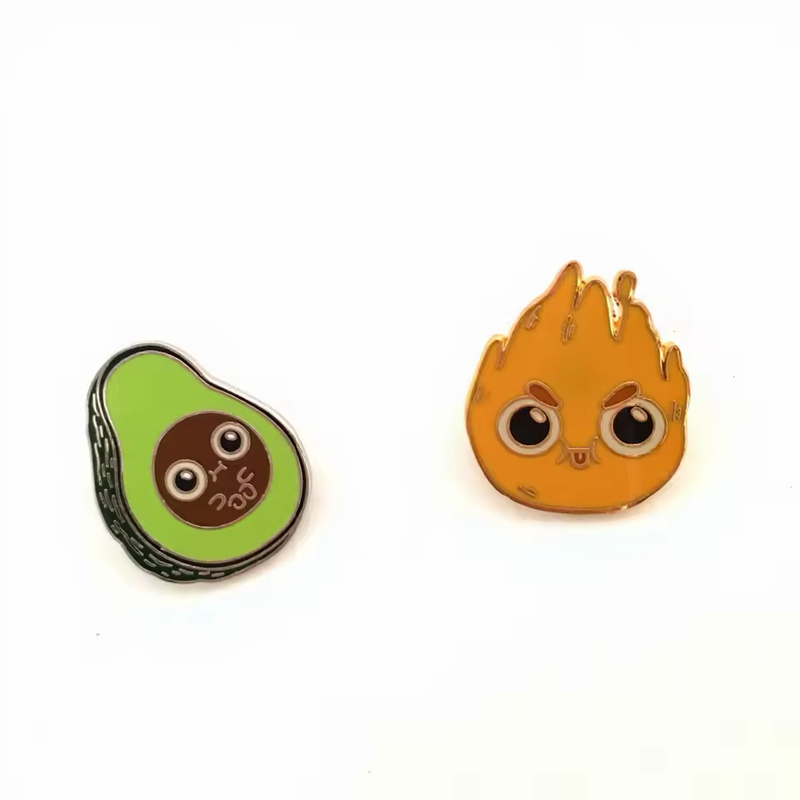 Green avocado and orange flame character pins with large round eyes displayed side by side on a white background.