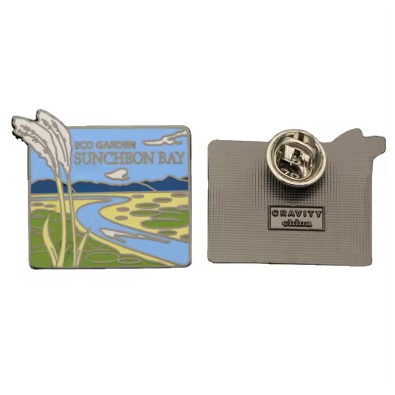 Scenic bay souvenir pin showing reeds, a winding blue waterway and distant hills, beside its textured metal back and butterfly clutch.