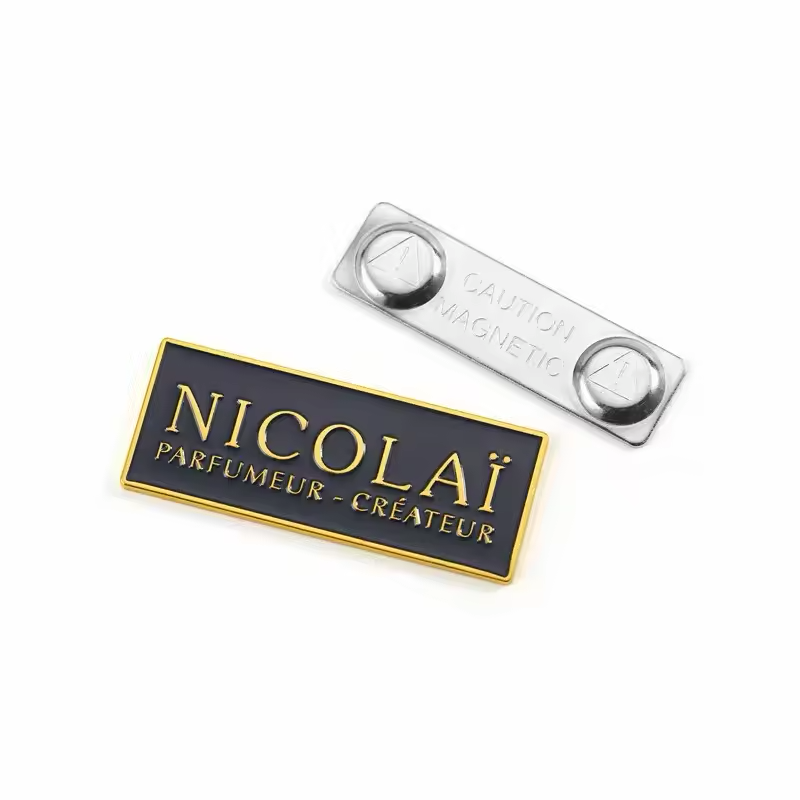 Black rectangular badge with gold-tone NICOLAI Parfumeur Createur lettering and border, beside a silver-tone two-disc fastener marked Caution Magnetic on white.