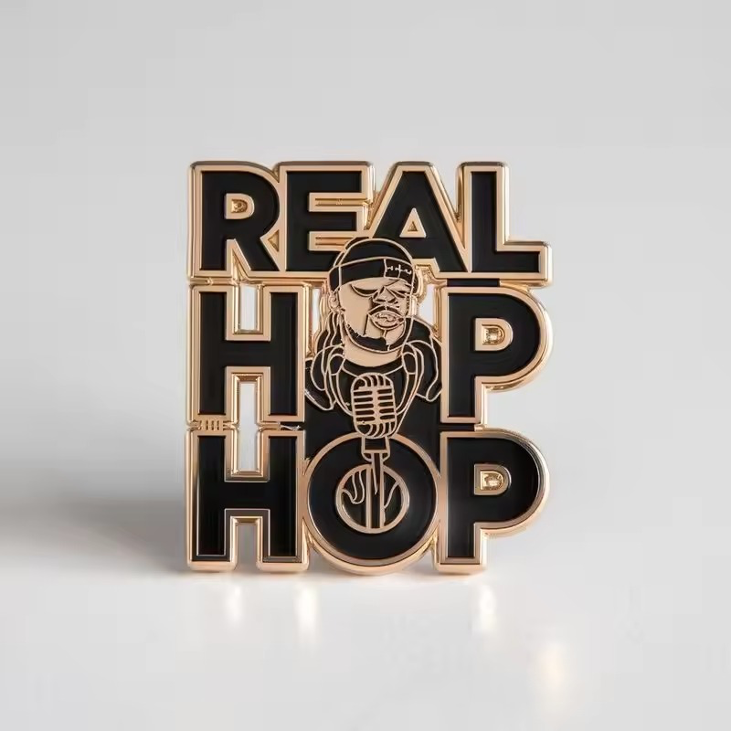 Black and gold-tone REAL HIP HOP pin with large stacked lettering, a central character portrait wearing a cap and sunglasses, and a microphone on a pale gray background.