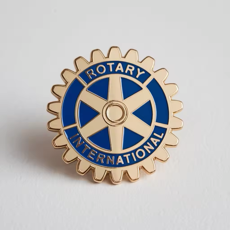 Gear-shaped Rotary emblem pin with blue sections, gold-tone spokes and outlines, and ROTARY INTERNATIONAL lettering on a pale gray background.