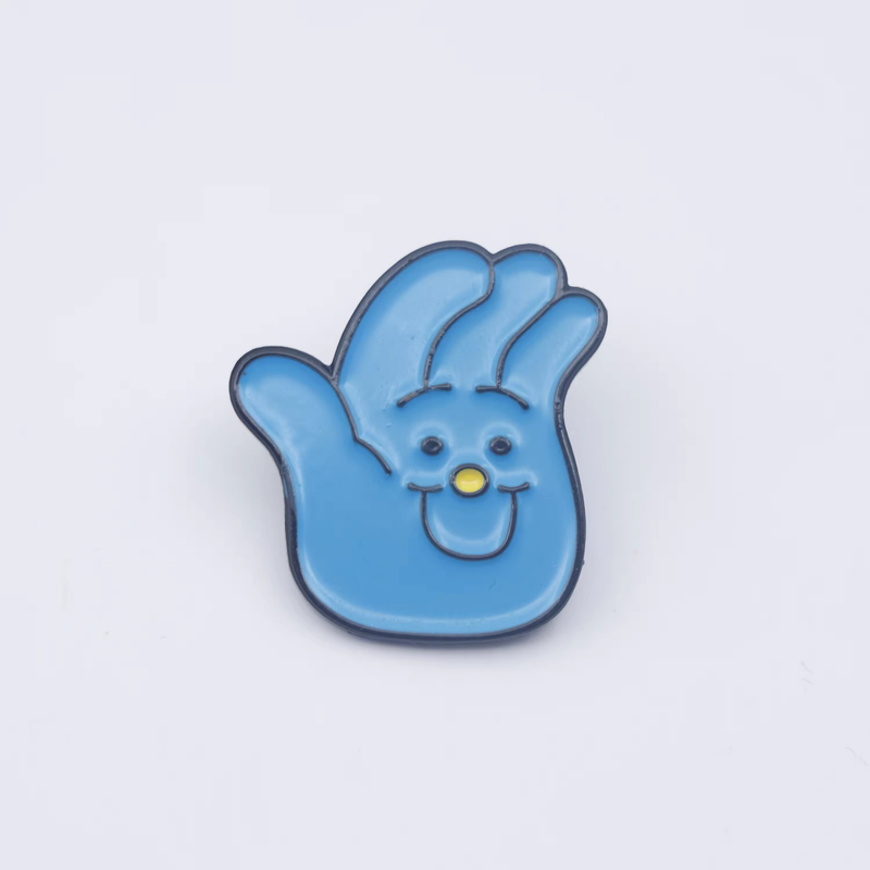 Blue hand-shaped cartoon pin with dark outlines, small dark eyes, a yellow round nose and a smiling mouth against a pale background.