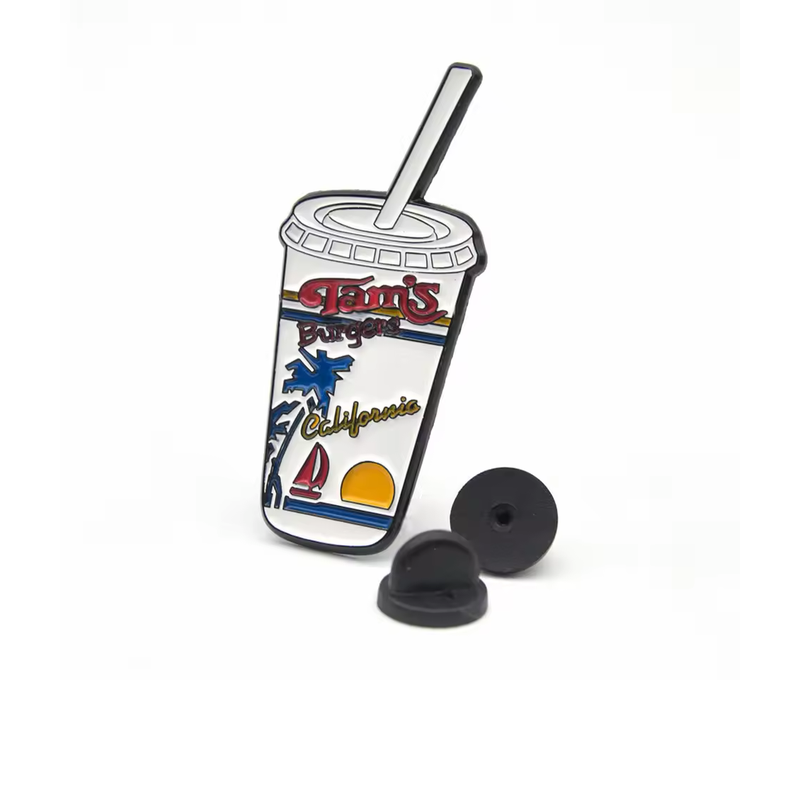 White drink cup pin with a straw, California lettering, palm trees, a sailboat and a sunset, shown with two black pin backs.