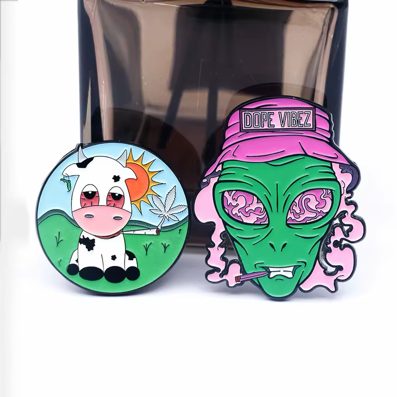 Cartoon pins showing a round cow scene with a sun, green hills, a serrated leaf and a smoking roll, beside a green alien wearing a pink DOPE VIBEZ hat with a smoking roll, in front of a dark display.