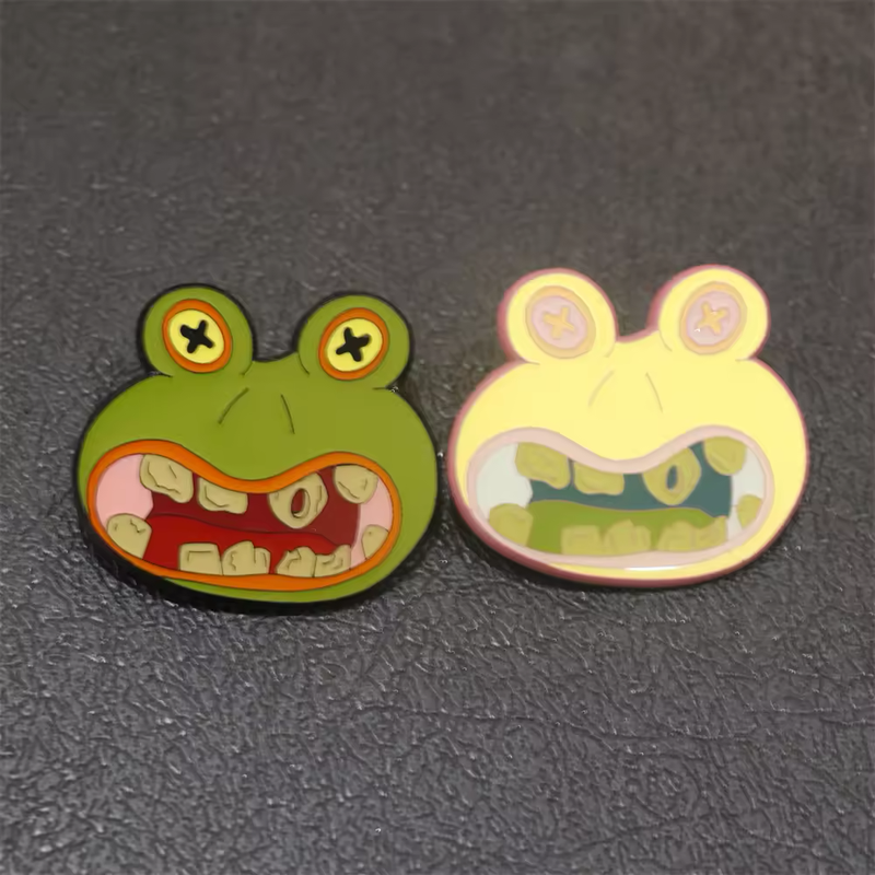 Green and pale yellow cartoon frog-face pins with X-shaped eyes and wide toothy mouths.