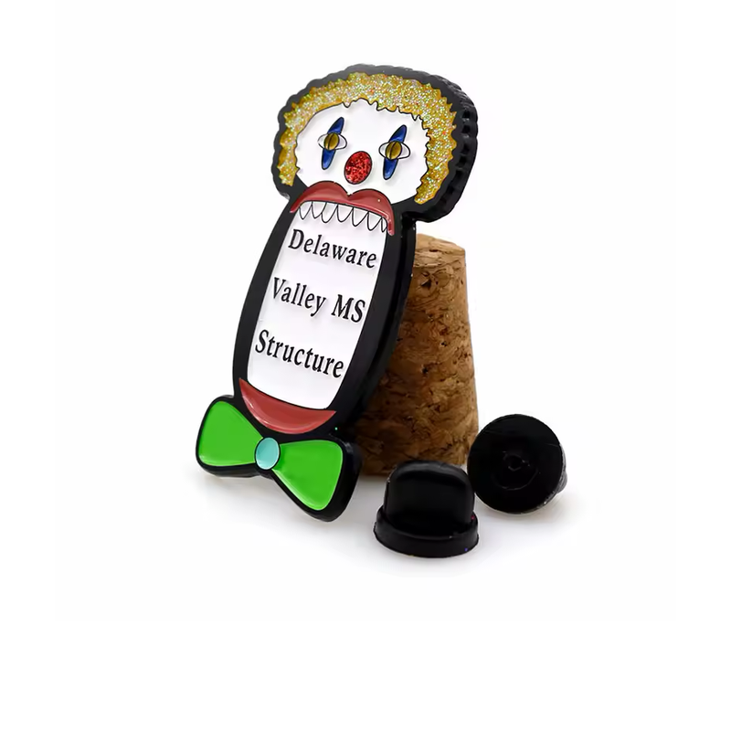 Clown-shaped pin with yellow glitter hair, a red glitter nose, a green bow tie and Delaware Valley MS Structure lettering, propped against cork beside two black rubber clutches.