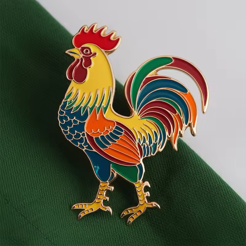 Colorful rooster pin with a red comb, yellow neck, multicolored feathers and gold-tone outlines on green fabric with a pale background.