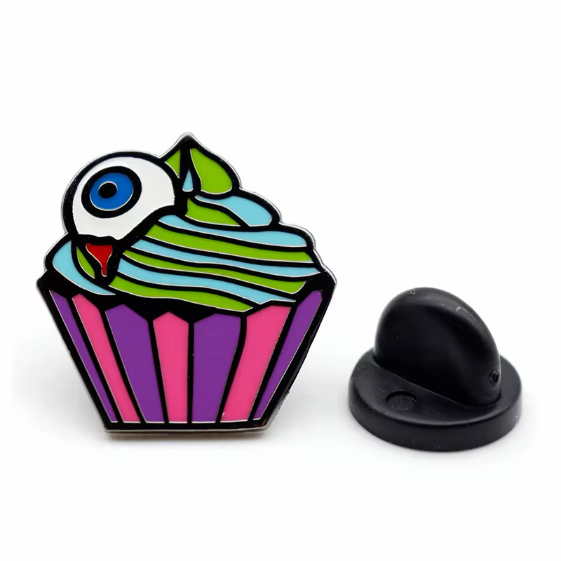 Cupcake pin with a blue eyeball, green and pale blue frosting and a purple and pink wrapper, shown beside a black rubber clutch.