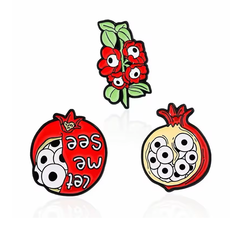 Three novelty pins featuring a red flower sprig and two pomegranates with eyeball motifs, including one with Let me see lettering.
