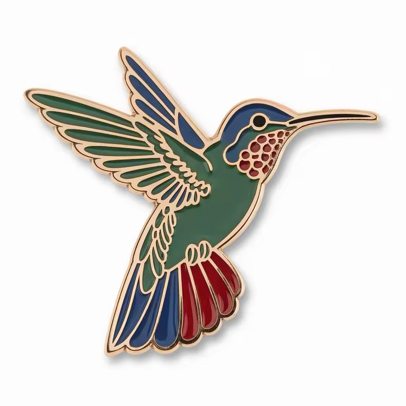 Flying hummingbird pin with a green body, blue head and wing details, red throat and tail accents, a long beak and gold-tone outlines on a white background.