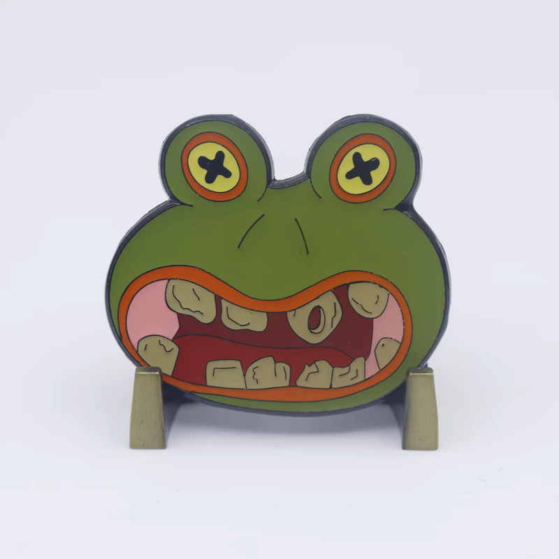 Close-up of a green frog-face pin with X-shaped eyes and a wide toothy mouth on a display stand.