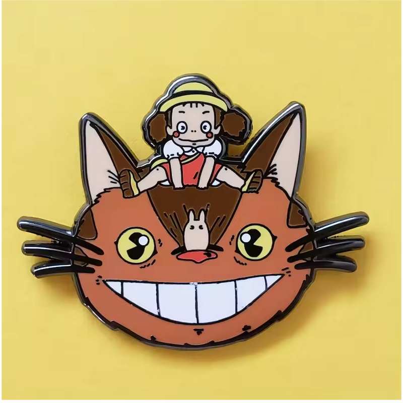 Cartoon pin showing a girl in a hat above a wide-grinning orange cat face, with yellow eyes, long whiskers and a small pale character on its nose, on a yellow background.