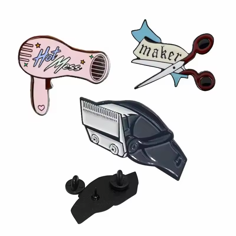 Barber tool pins showing a pink Hot Mess hair dryer, scissors with a maker banner, and a black and silver-tone hair clipper, with a reverse view and black rubber clutches on white.