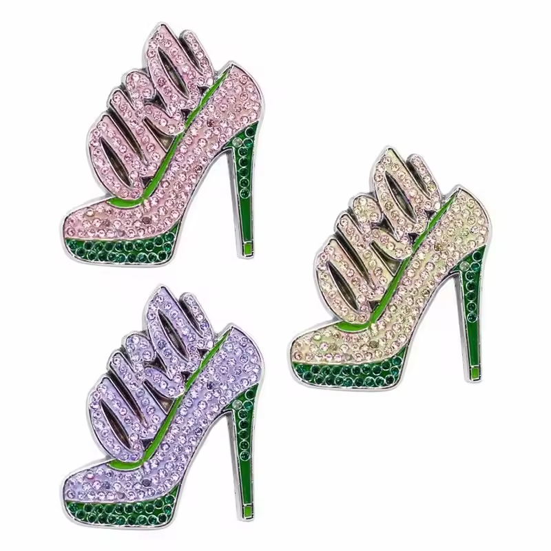 Three rhinestone high heel shoe brooches with large letter decorations in pink, pale and purple colors, with green soles and heels on a white background.