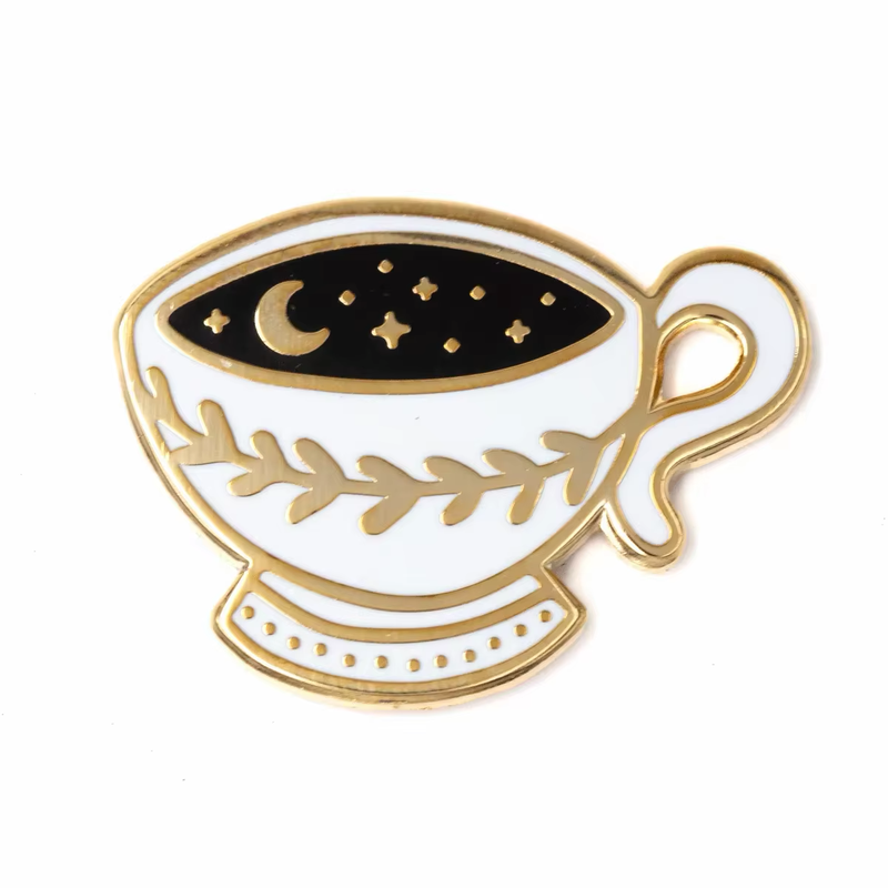 White teacup pin with gold-tone edging and a leafy branch, with a crescent moon and stars in the black drink.