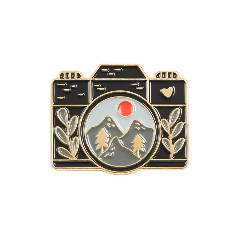 Black camera-shaped pin with mountains, pine trees and a red sun inside the lens, framed by leafy details.