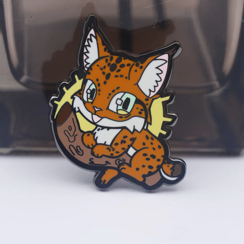 Orange spotted cartoon cat pin with tufted ears, pale green eyes, a white muzzle and chest, sitting on a brown branch with yellow accents against a dark display background.