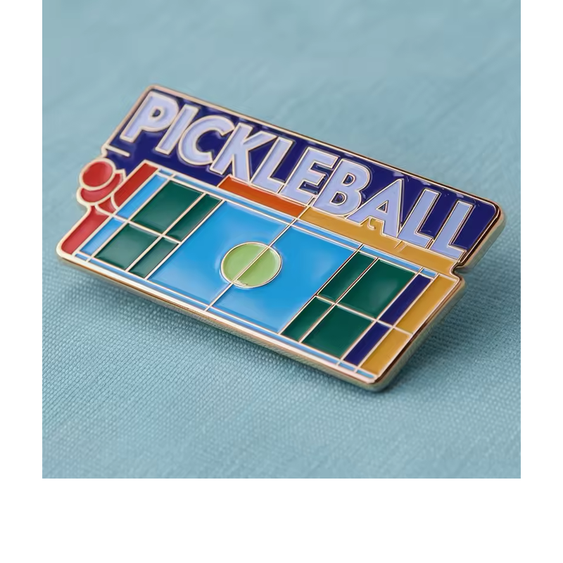 Pickleball court-shaped pin with blue and green court sections, white boundary lines, PICKLEBALL lettering and gold-tone outlines on a blue fabric background.