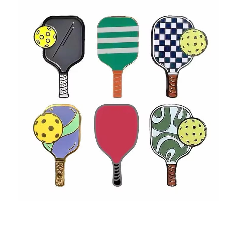 Six pickleball paddle pins with solid, striped, checkered and abstract patterns, including four designs paired with yellow perforated balls.