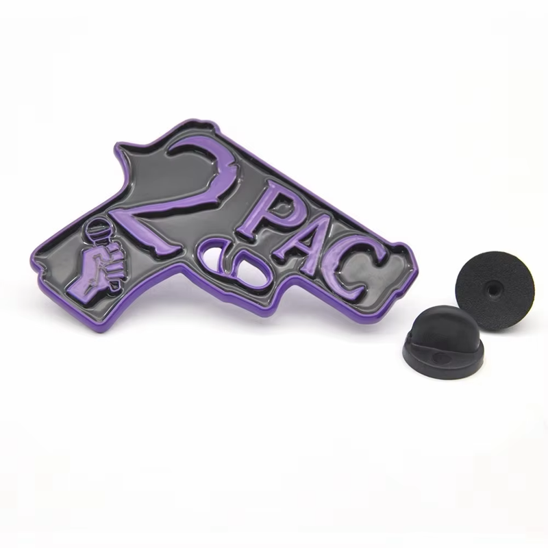 Gray pistol-shaped pin with purple 2PAC lettering and a hand holding a microphone, beside two black rubber pin clutches on a white background.