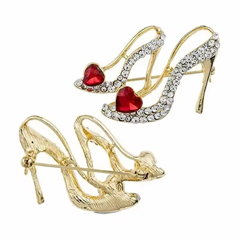 Gold-tone high heel shoe brooch with clear rhinestones and red heart-shaped stones, shown from the front and back with its pin clasp on a white background.
