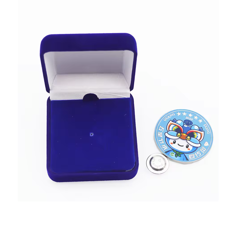 Round light blue mascot pin with a colorful character, stars and lettering beside an open blue presentation box and a silver-tone butterfly clutch on a white background.