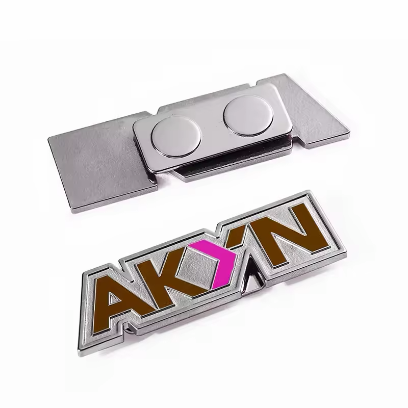 Silver-tone shaped logo badge with brown lettering and a pink geometric accent, shown front and back with a rectangular two-disc magnetic fastener on a white background.