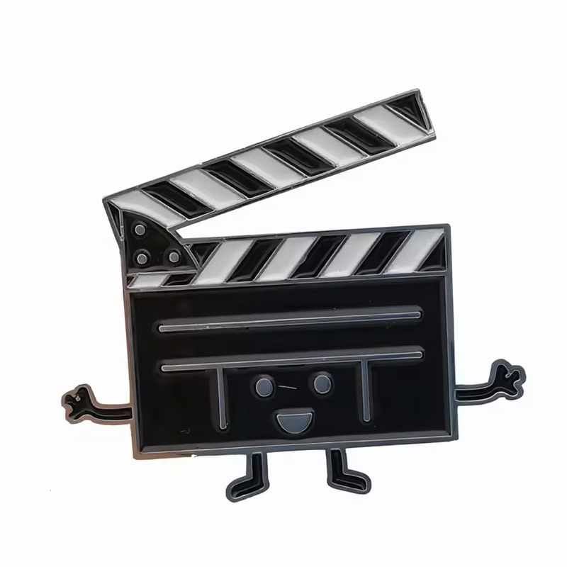 Black and white film clapperboard character pin with an open striped clapstick, smiling face, outstretched arms and small legs on a white background.
