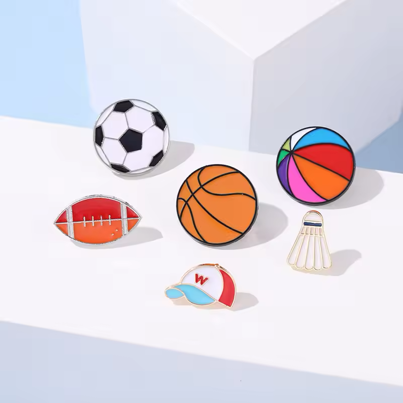 Six sports pins showing a soccer ball, an orange basketball, a multicolored ball, an American football, a shuttlecock and a cap on white display blocks.