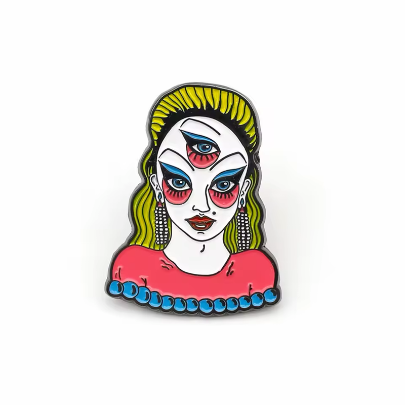 Three-eyed portrait pin with yellow-green hair, blue eye makeup, pink cheeks, dangling earrings and a pink top edged with blue beads on a white background.