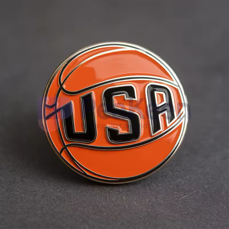 Orange basketball pin with large black USA lettering and silver-tone outlines on a dark textured background.