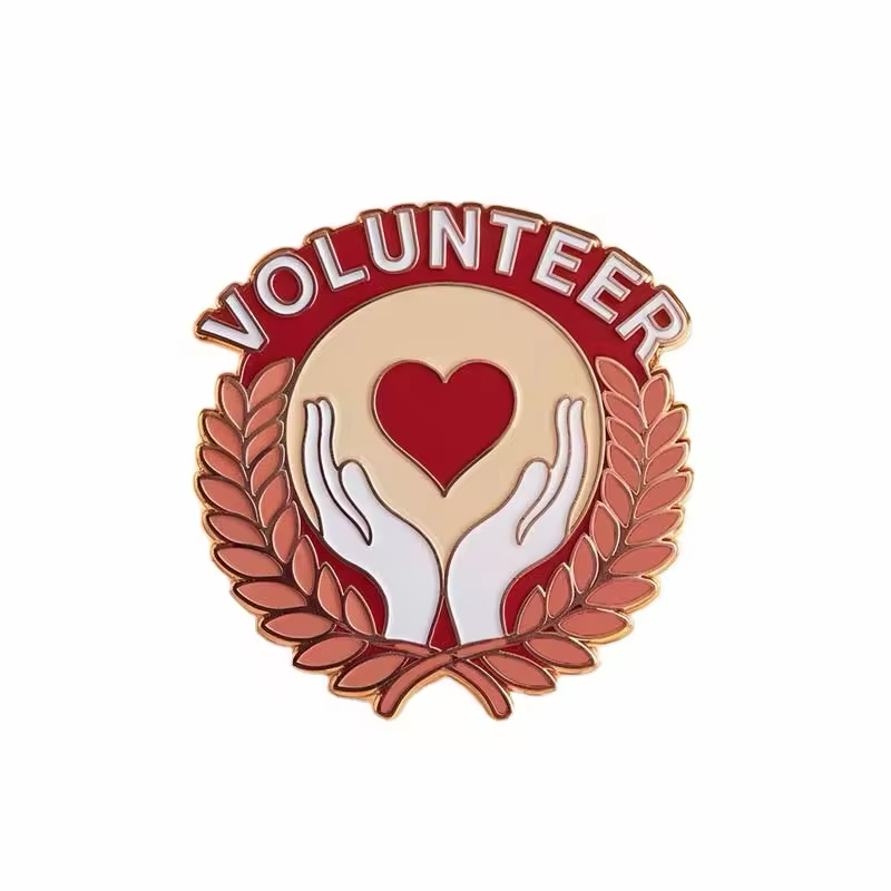 Volunteer pin with white VOLUNTEER lettering on a red banner, a red heart above two white hands, warm-toned laurel branches and gold-tone outlines on a white background.