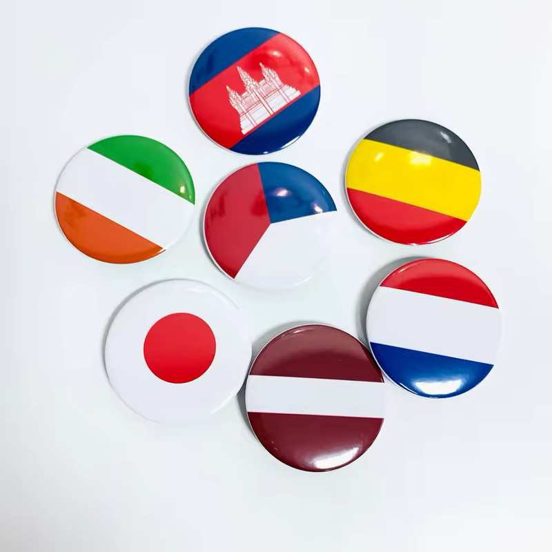 Seven glossy round flag-themed button badges, including Cambodia, Ireland, Belgium, Japan, the Netherlands and Latvia designs and a red-white-blue design, on a pale surface.