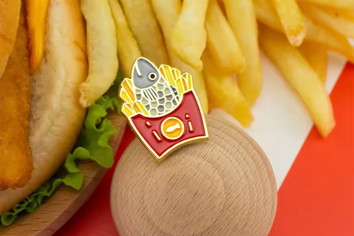 A red fries-shaped enamel pin displayed on a wooden stand beside a burger and fries.