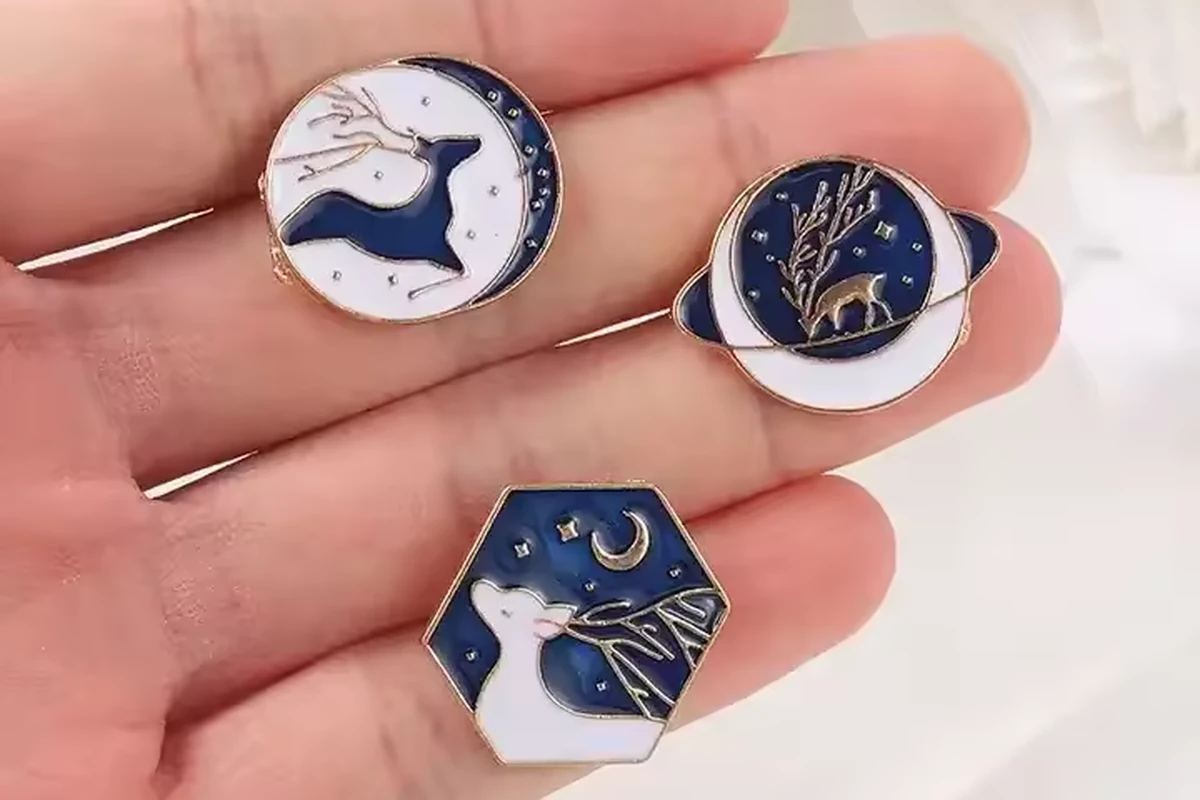 A hand holding three blue and white deer-design pins in round, ringed and hexagonal shapes.